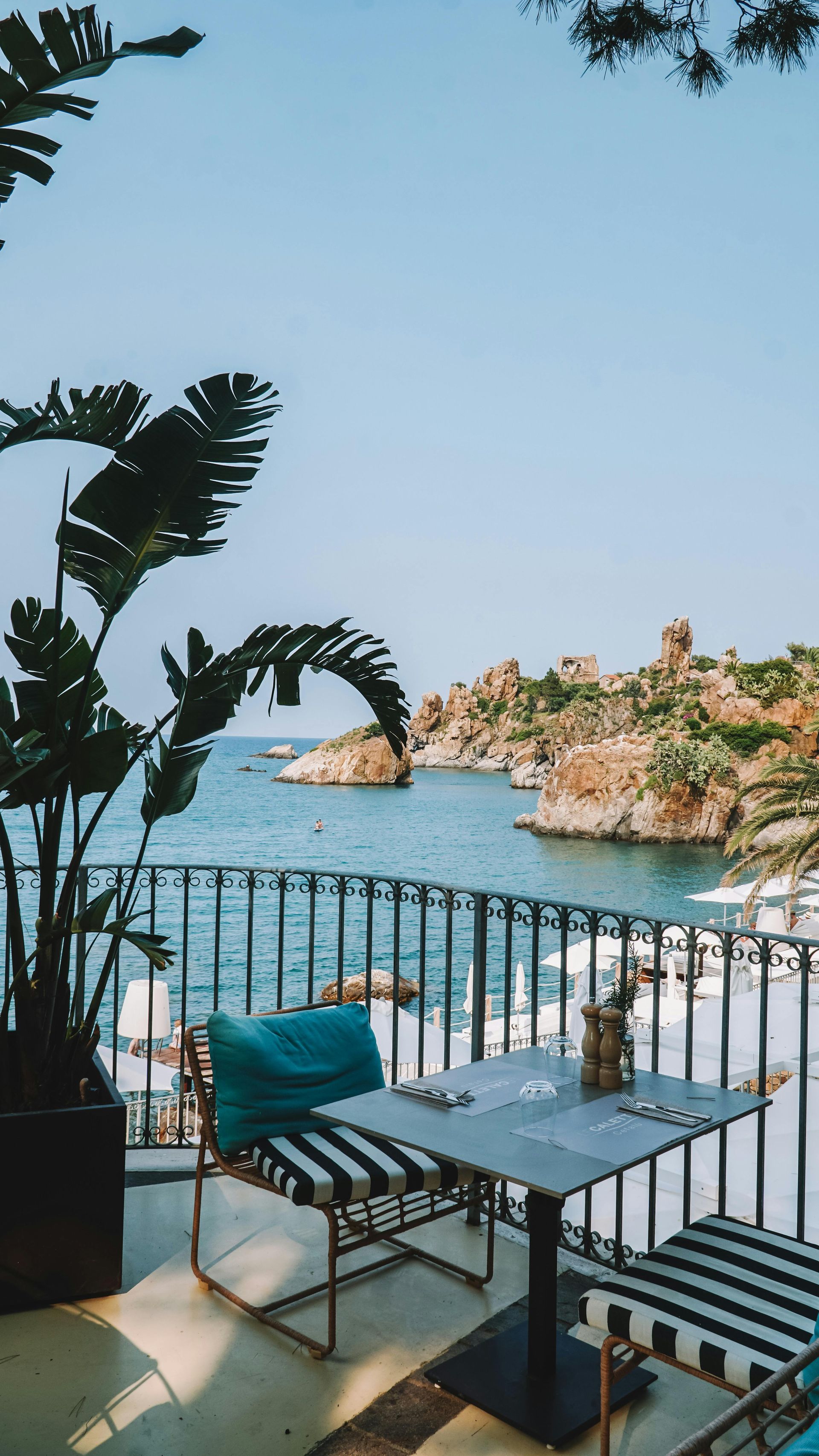 Balcony with table and chairs overlooking the sea and rocky islands under a blue sky.