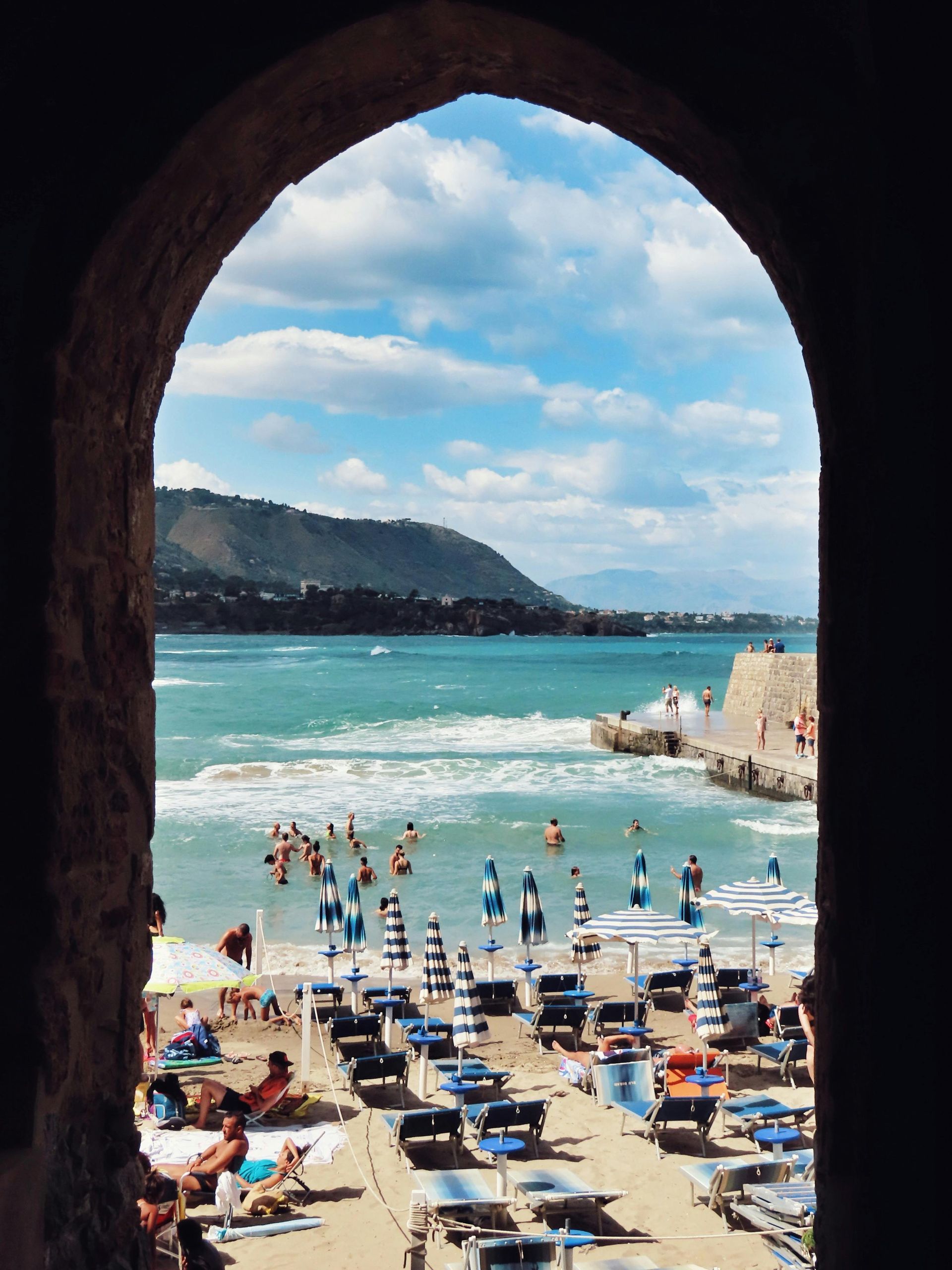 Beach scene viewed through an archway: People swim and relax on beach with blue sky, sea, and mountains.