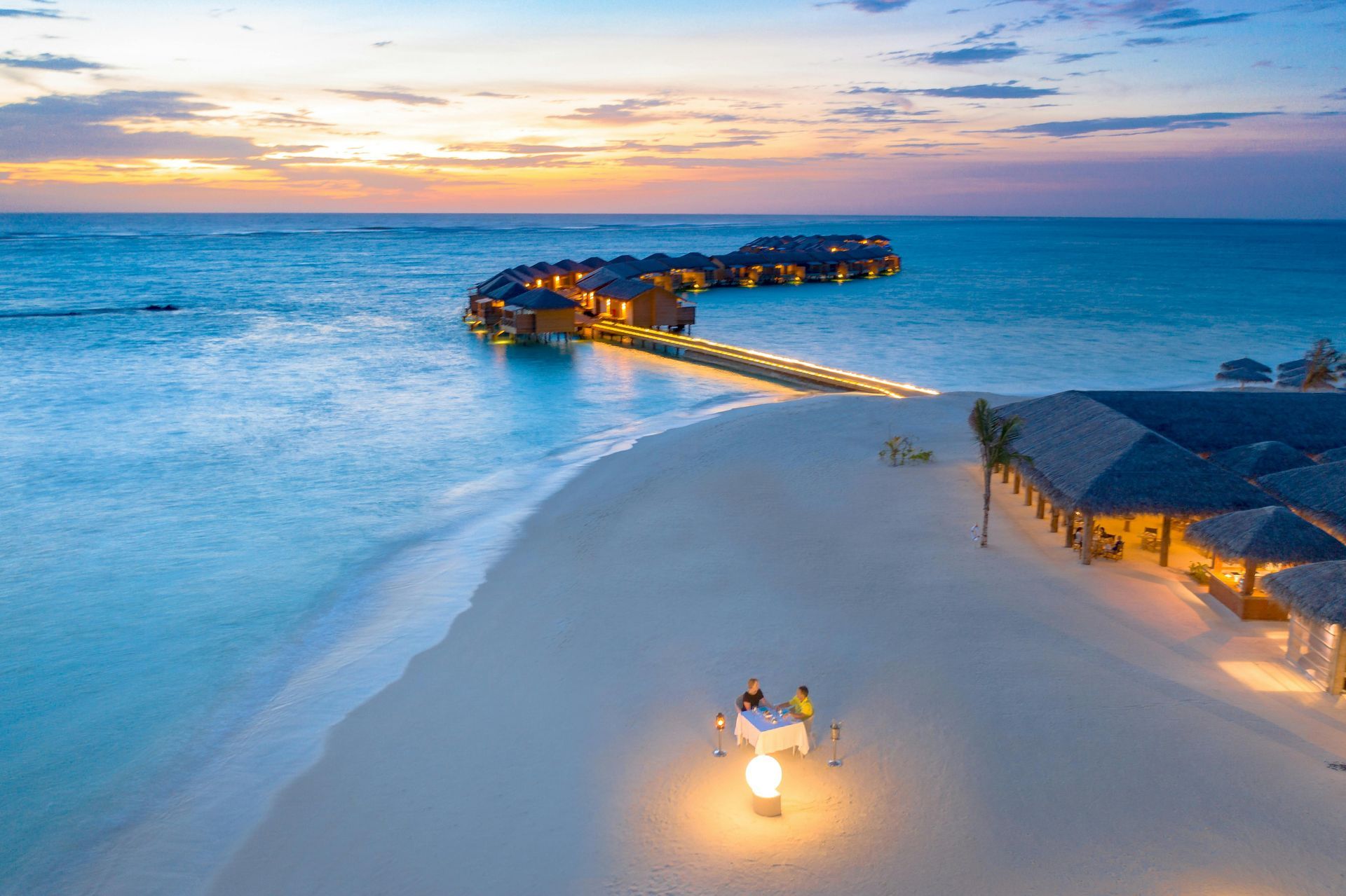 Beachfront resort with overwater bungalows, calm turquoise water, and a couple dining at sunset.