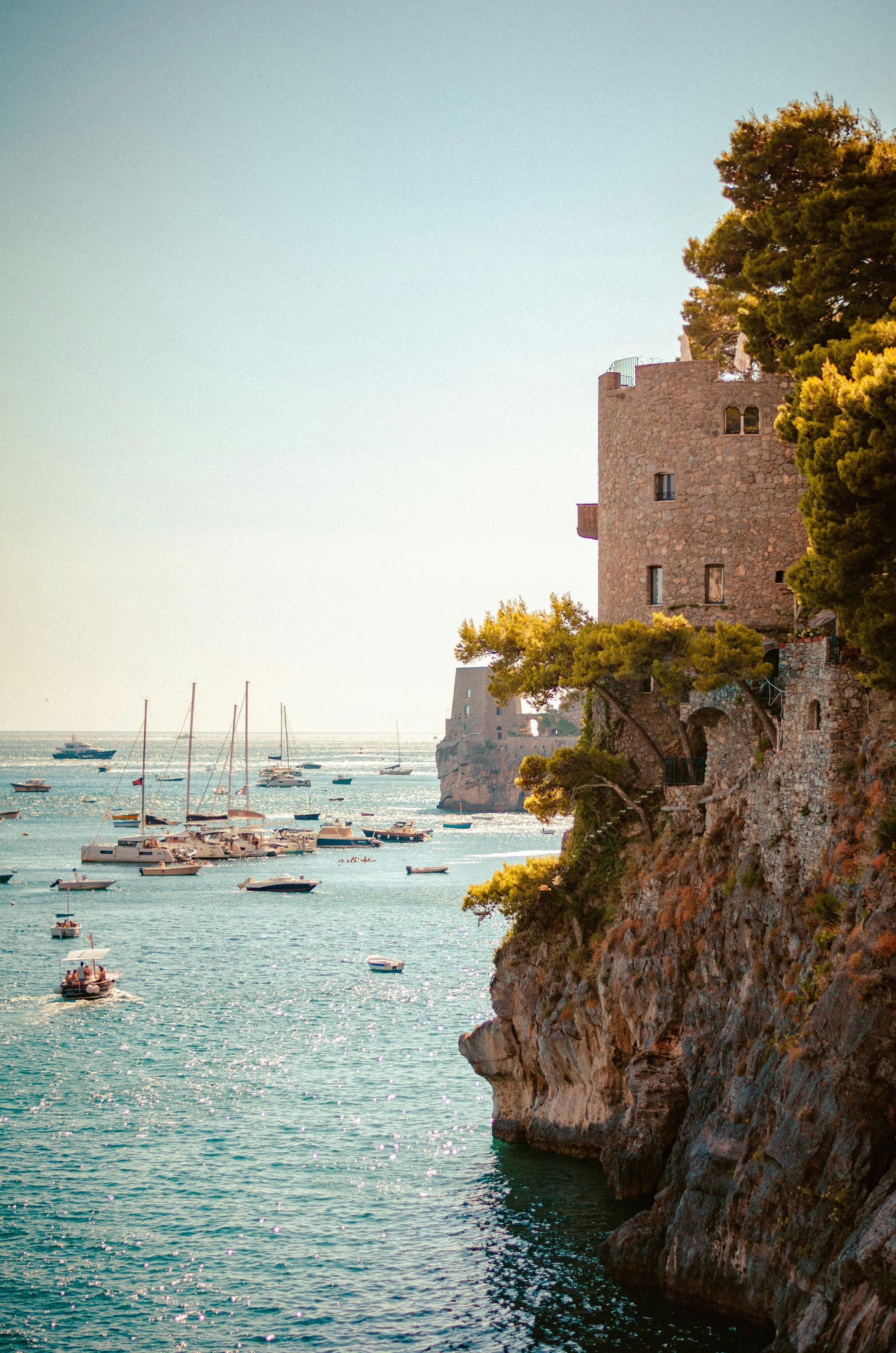 Stone tower perched on a rocky cliff overlooking a bay filled with sailboats on a sunny day.