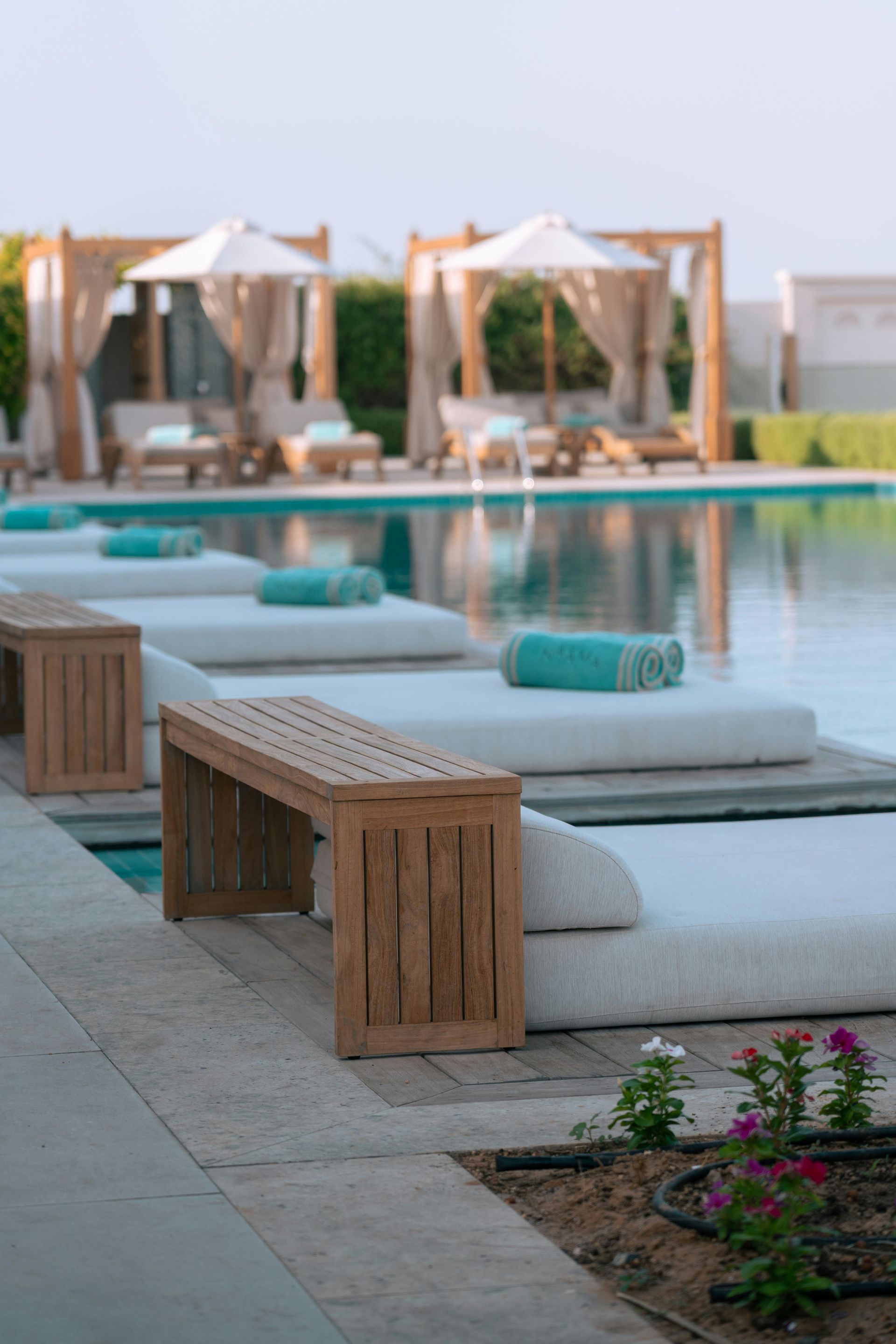 Poolside scene with lounge chairs, turquoise towels, wooden benches, and cabanas.