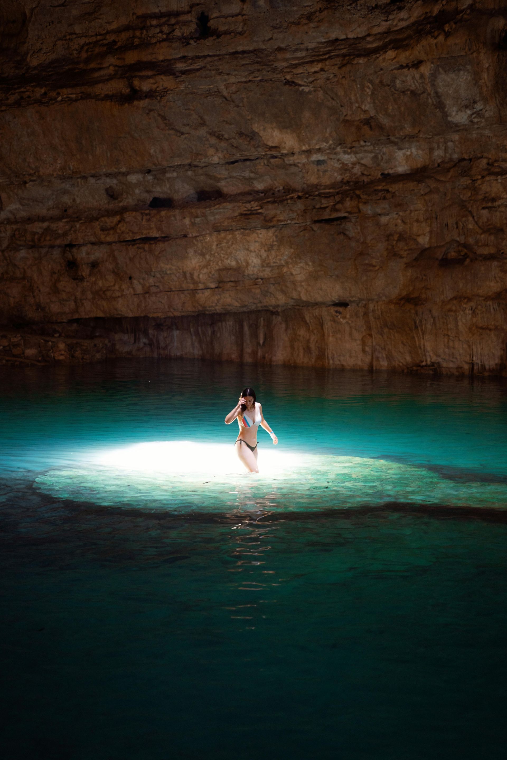 Woman in a swimsuit stands in the turquoise water of a cave pool, with light shining down.