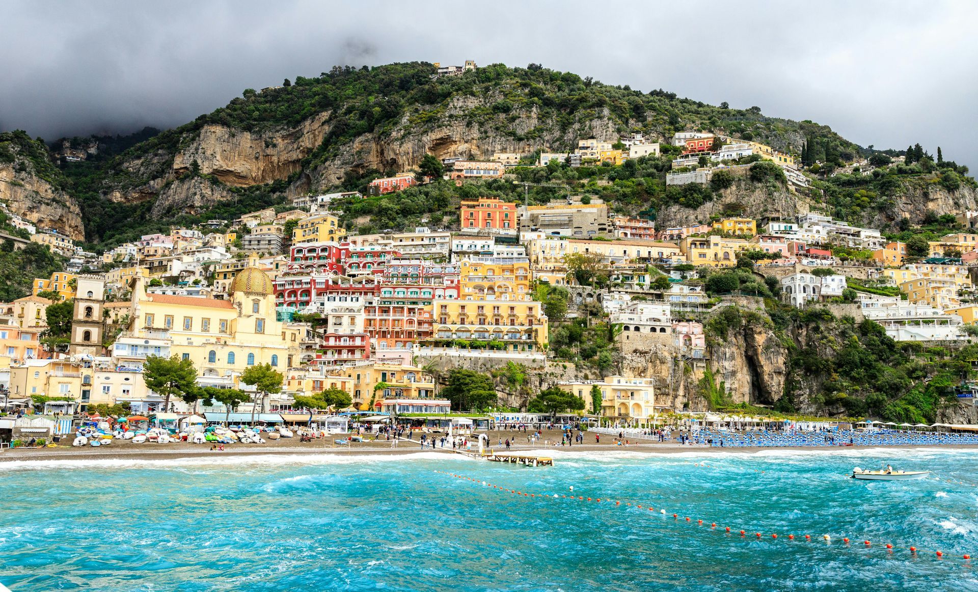 A seaside town with colorful buildings stacked on a steep cliffside above a turquoise beach under a cloudy sky.