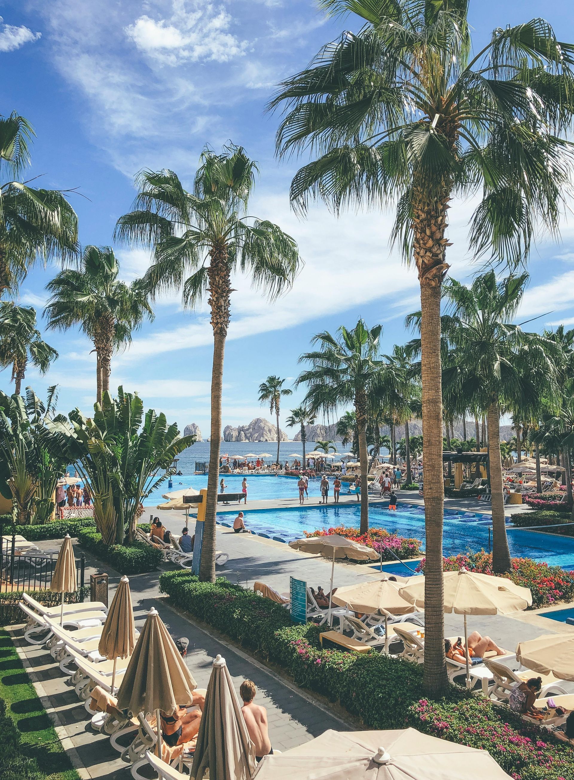 A sunny resort pool area with palm trees, lounge chairs with umbrellas, and people swimming under a bright blue sky.