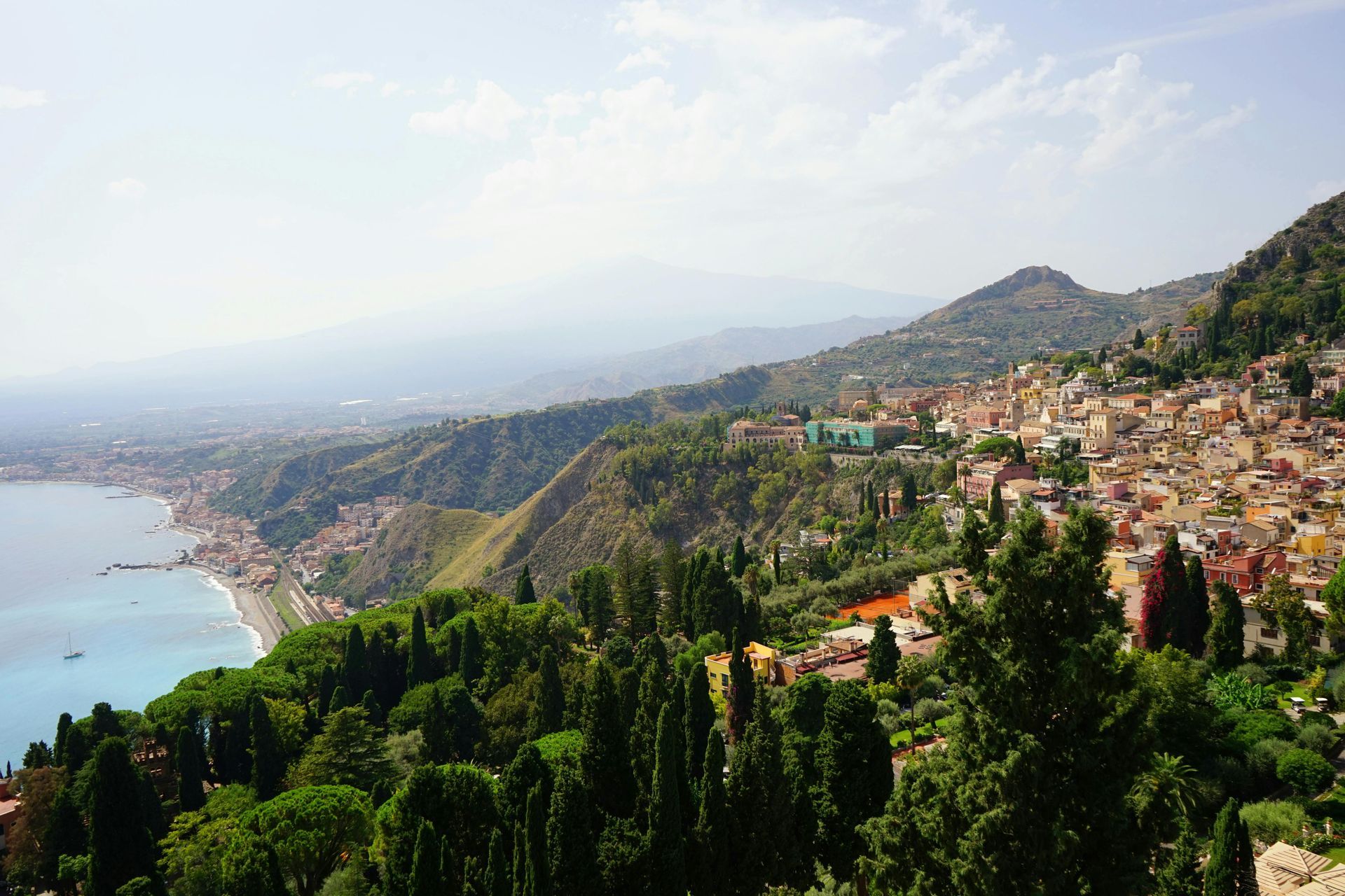 Coastal Sicilian town, sea on the left, hillside homes, green trees, mountains in the distance, blue sky.