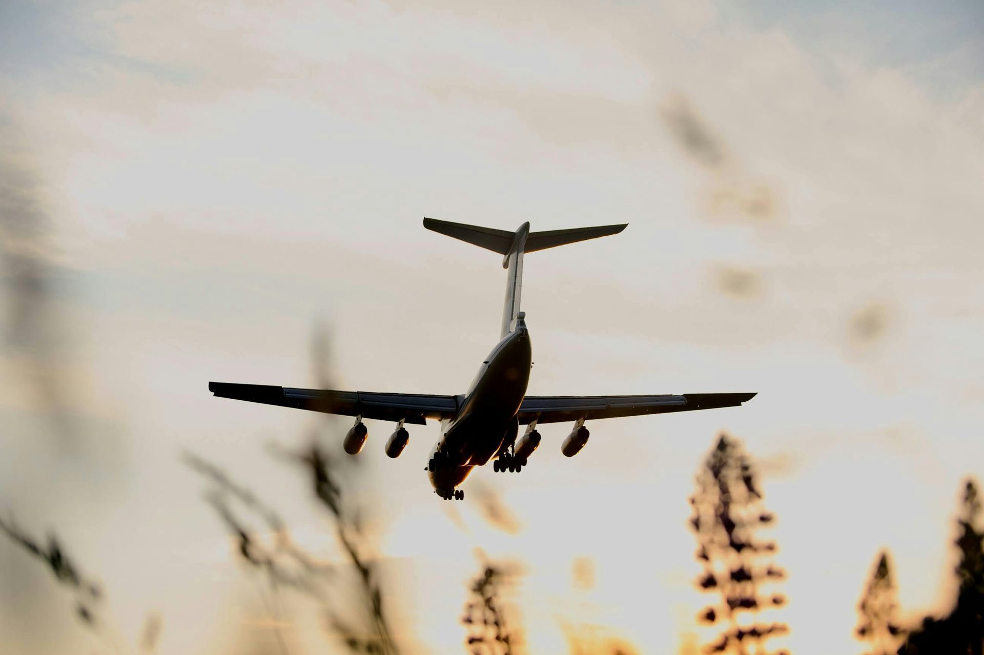 A silhouette of a four-engine airplane flying low against a soft, golden-hued sky above silhouetted trees.