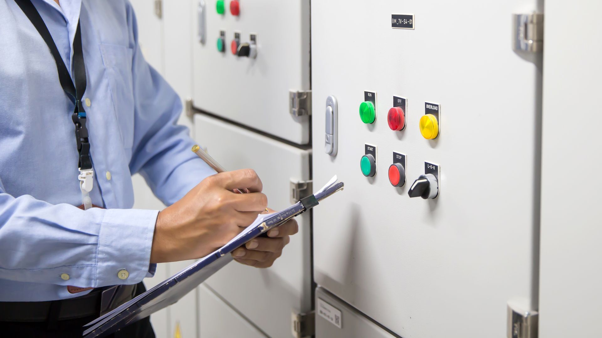 A man is holding a clipboard and writing on it in front of a control panel.