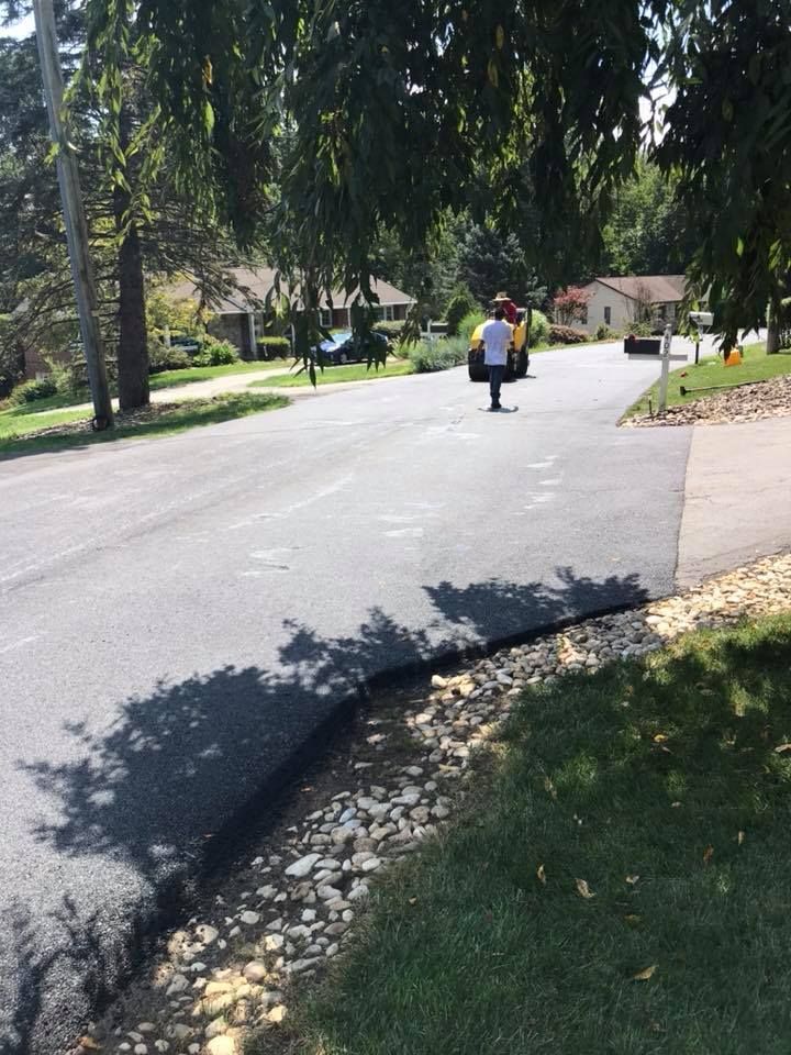 Person stands on newly paved road. Asphalt is black, bordered by grass and rocks. Sun shines.