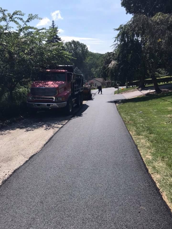 Red dump truck on a freshly paved road, worker shoveling asphalt, trees line sides, sunny day.