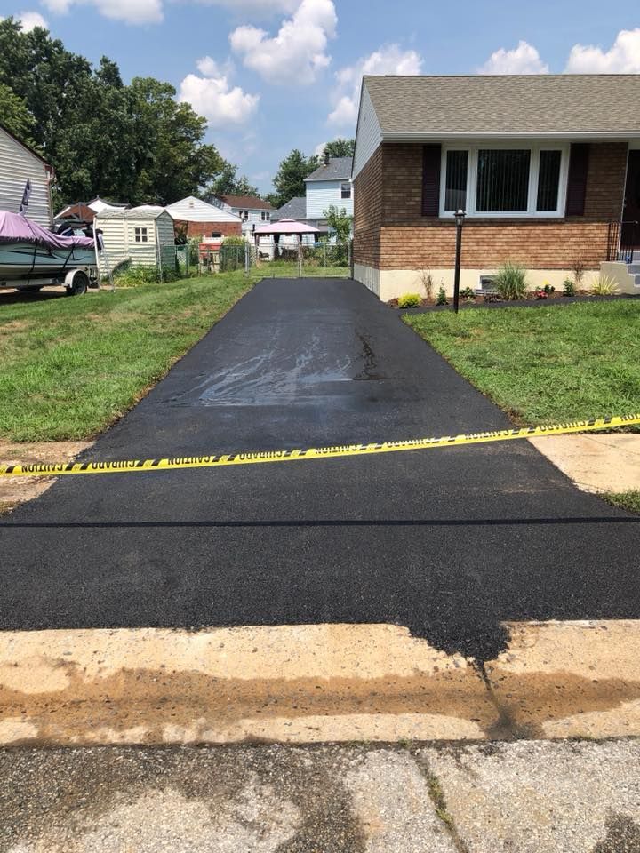 Newly paved asphalt driveway, with yellow caution tape, leading to a brick house on a sunny day.