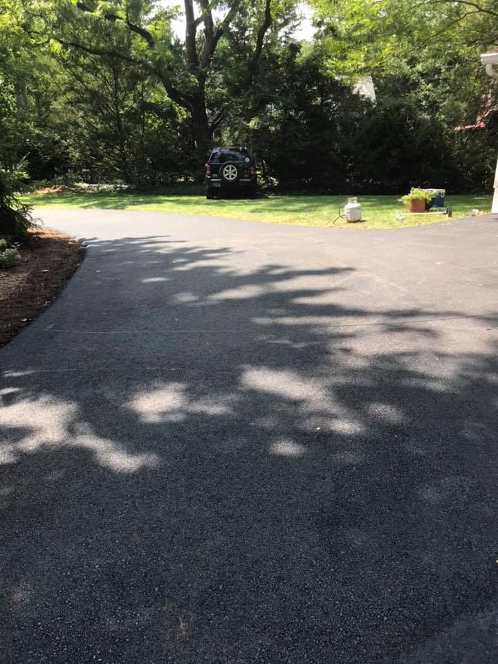 Paved driveway leads into a tree-lined yard with a vehicle parked in the distance.