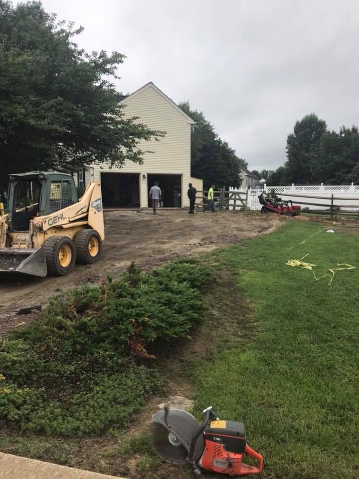 Construction scene: Skid steer, workers, and a saw near a garage with open doors and a yard.