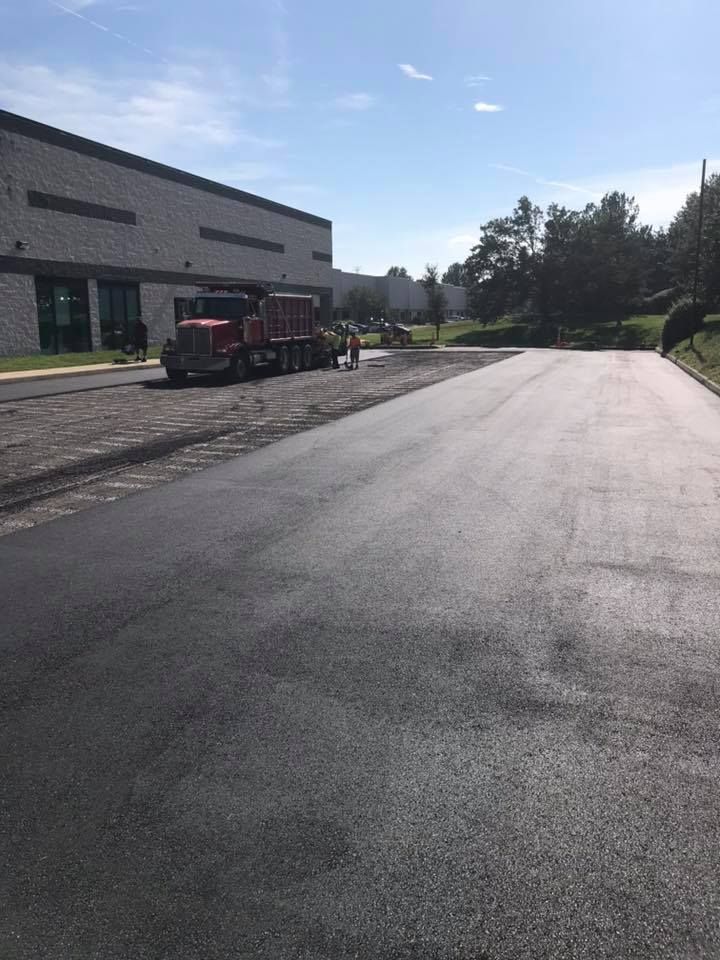 Asphalt road paving near a large industrial building. A dump truck and workers are present.
