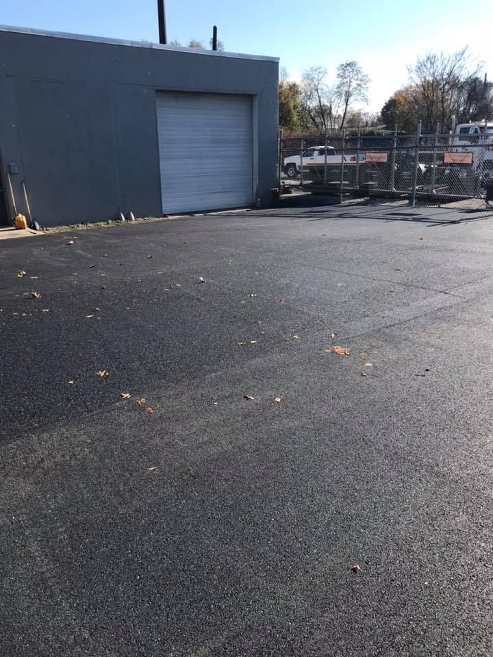 Asphalt parking lot next to a gray building with a closed garage door. Fencing and vehicles are visible in the background.