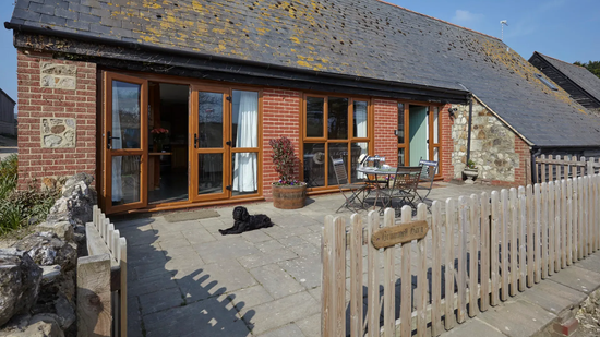 A rustic brick cottage with a sunny stone patio, a small wooden fence, and a black dog lying on the ground.