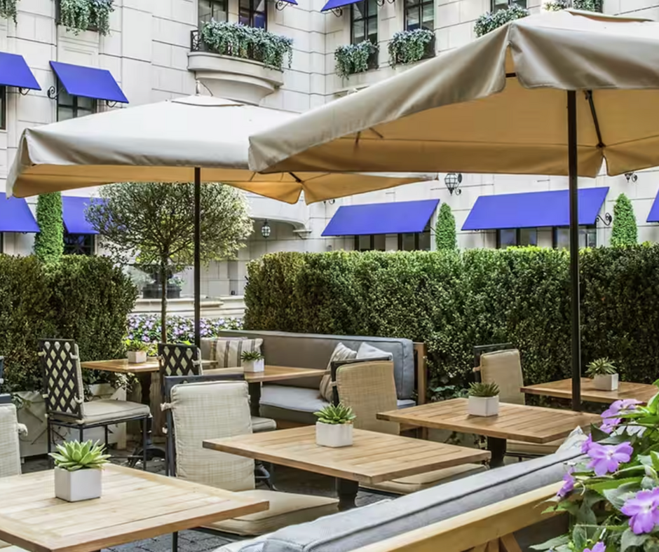 An outdoor café courtyard with wooden tables, beige umbrellas, green hedges, and blue window awnings.