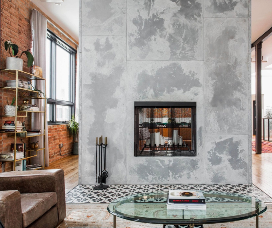 A modern living room features a large, gray concrete fireplace, a glass coffee table, and an exposed brick wall with shelves.
