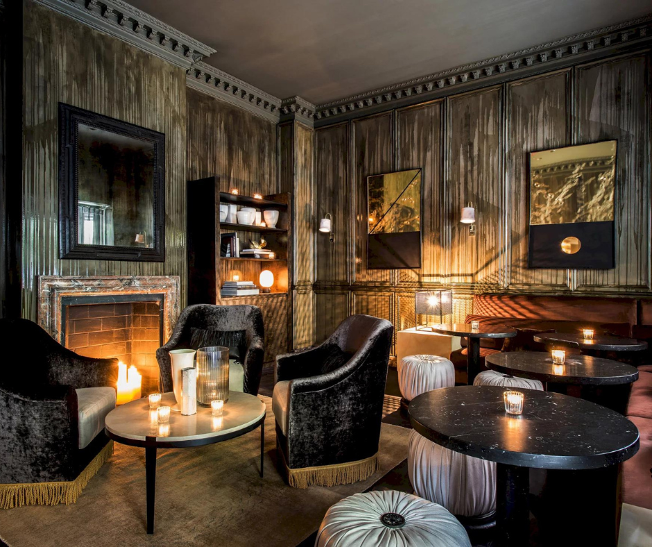 A cozy, dimly lit lounge featuring a stone fireplace, plush velvet chairs, circular tables, and wood-paneled walls.