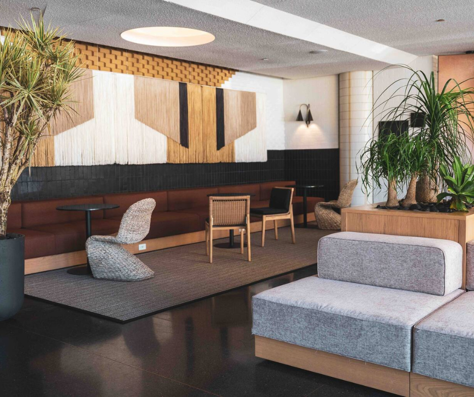 A modern lounge area with brown banquette seating, patterned chairs, decorative wall art, and several indoor plants.