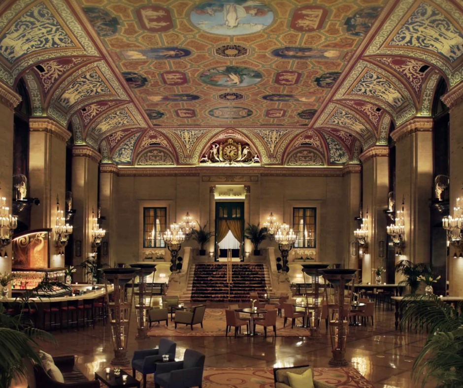 Grand hotel lobby with a vaulted, ornately painted ceiling, marble columns, elegant chandeliers, and plush seating.