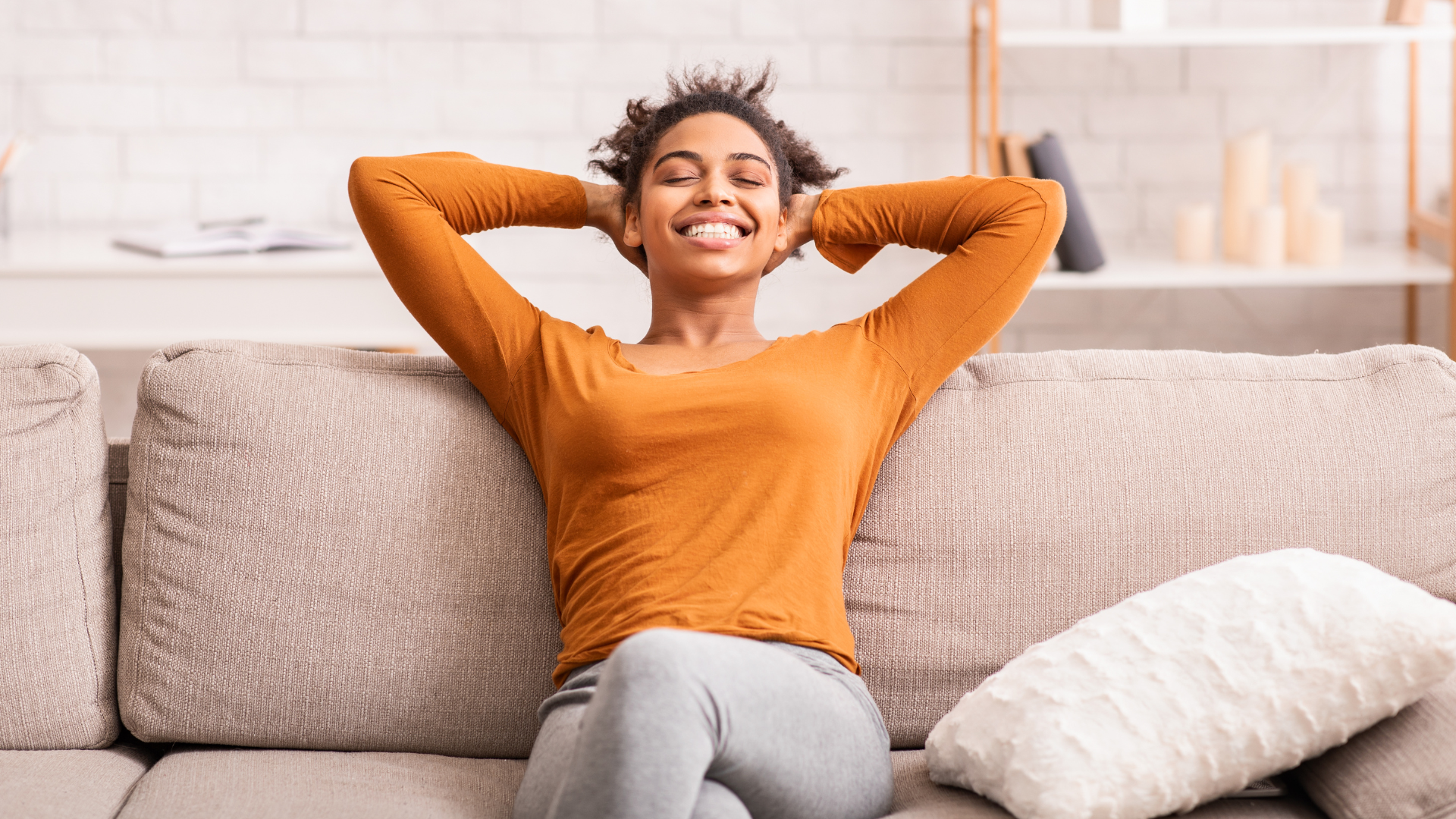 Person relaxing on a beige couch, smiling with hands behind head, wearing an orange sweater.