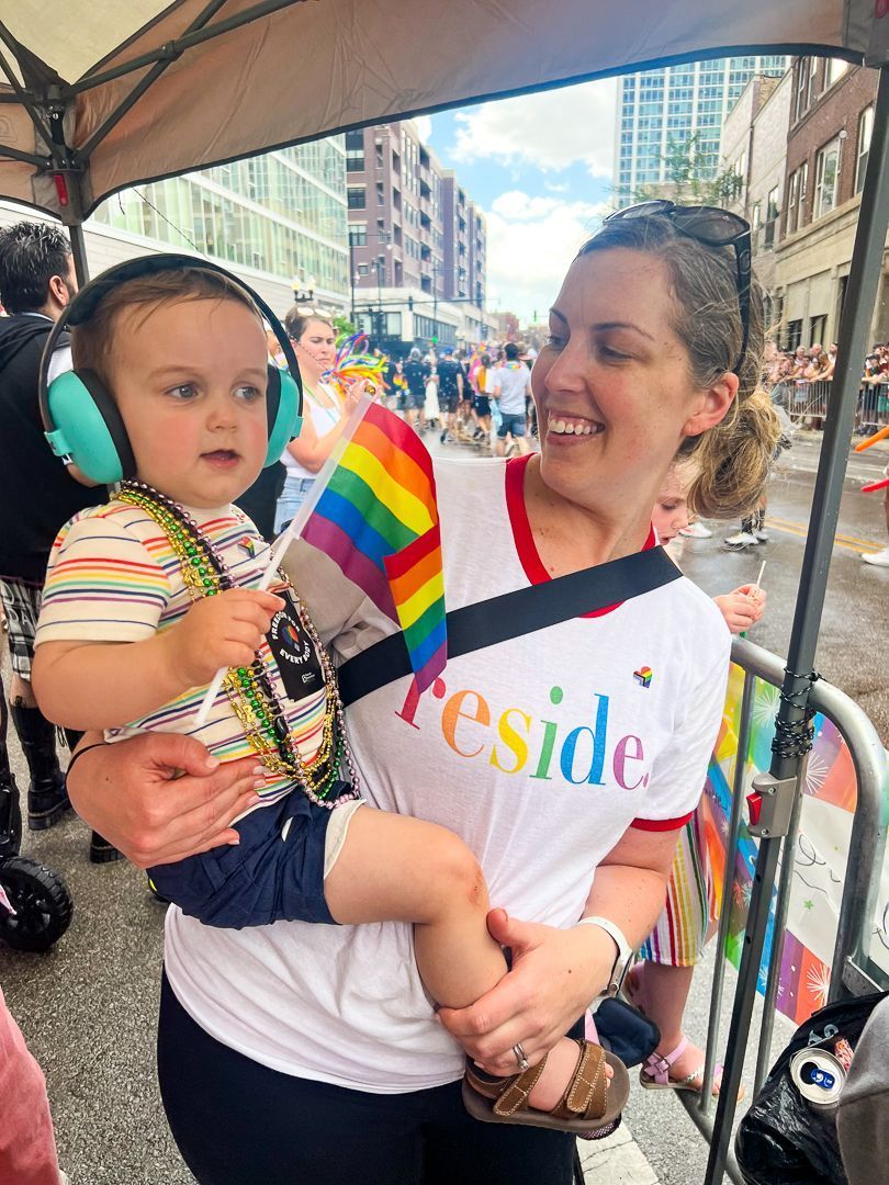 A woman is holding a baby wearing headphones and a rainbow flag.
