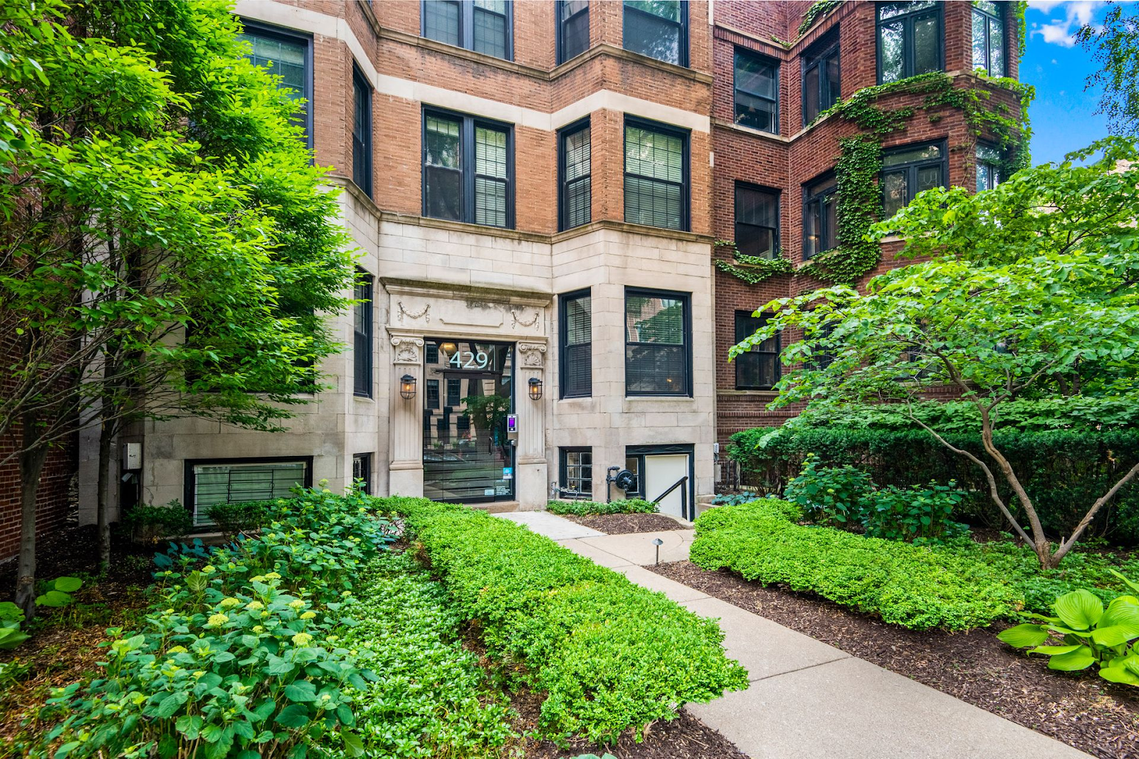Apartment building exterior with a paved path, lush greenery, and a decorative entrance.