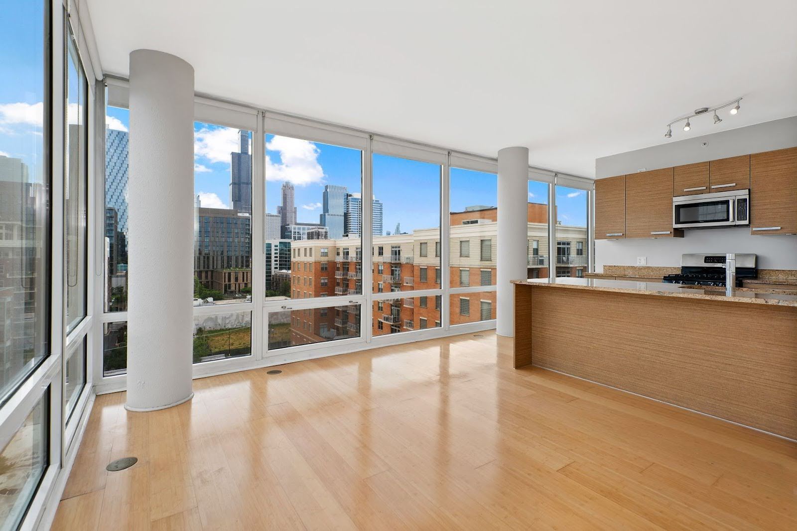 Modern apartment living area with light wood floors, floor-to-ceiling windows, cityscape view, and a kitchen island.