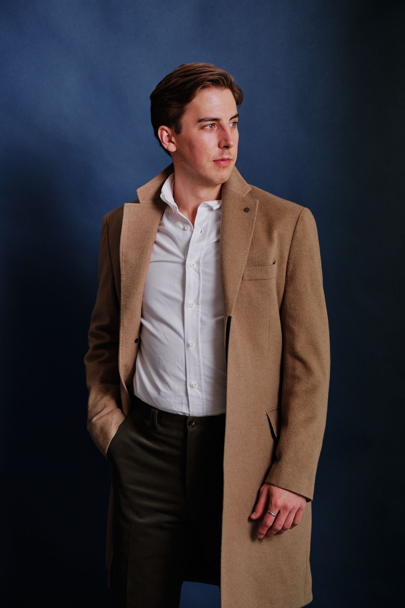 A man wearing a tan coat and a white shirt is standing with his hands in his pockets.