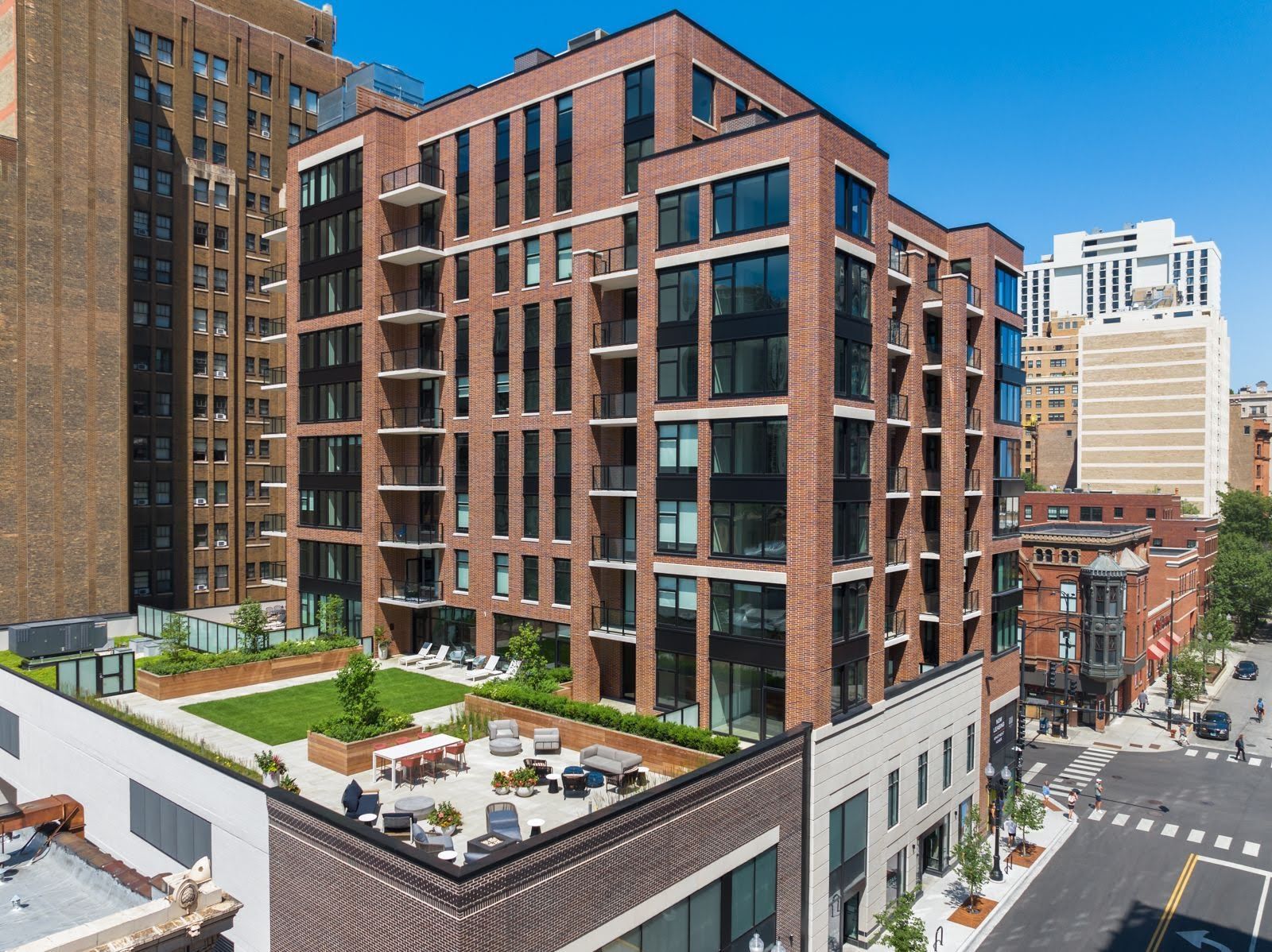 A modern brick apartment building in a city features an outdoor terrace with seating and lawn areas on the lower roof.
