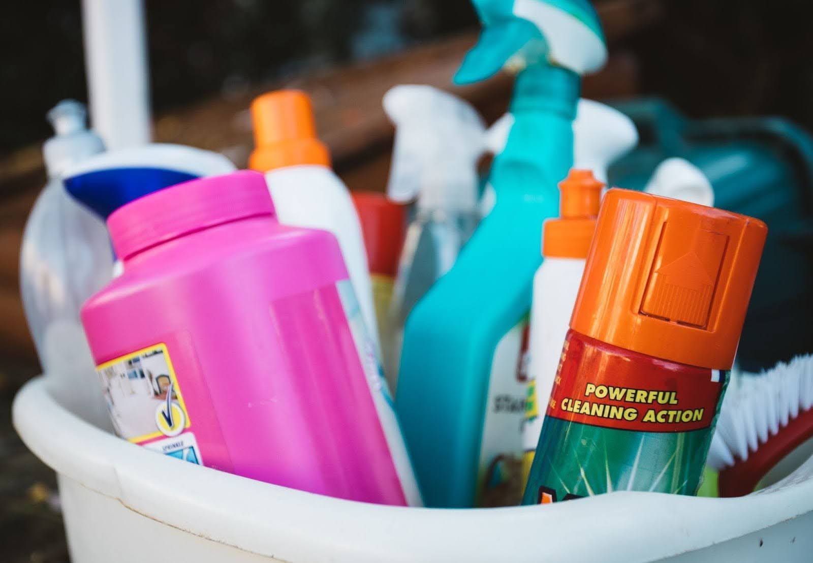 A white bucket filled with various brightly colored household cleaning bottles and brushes.