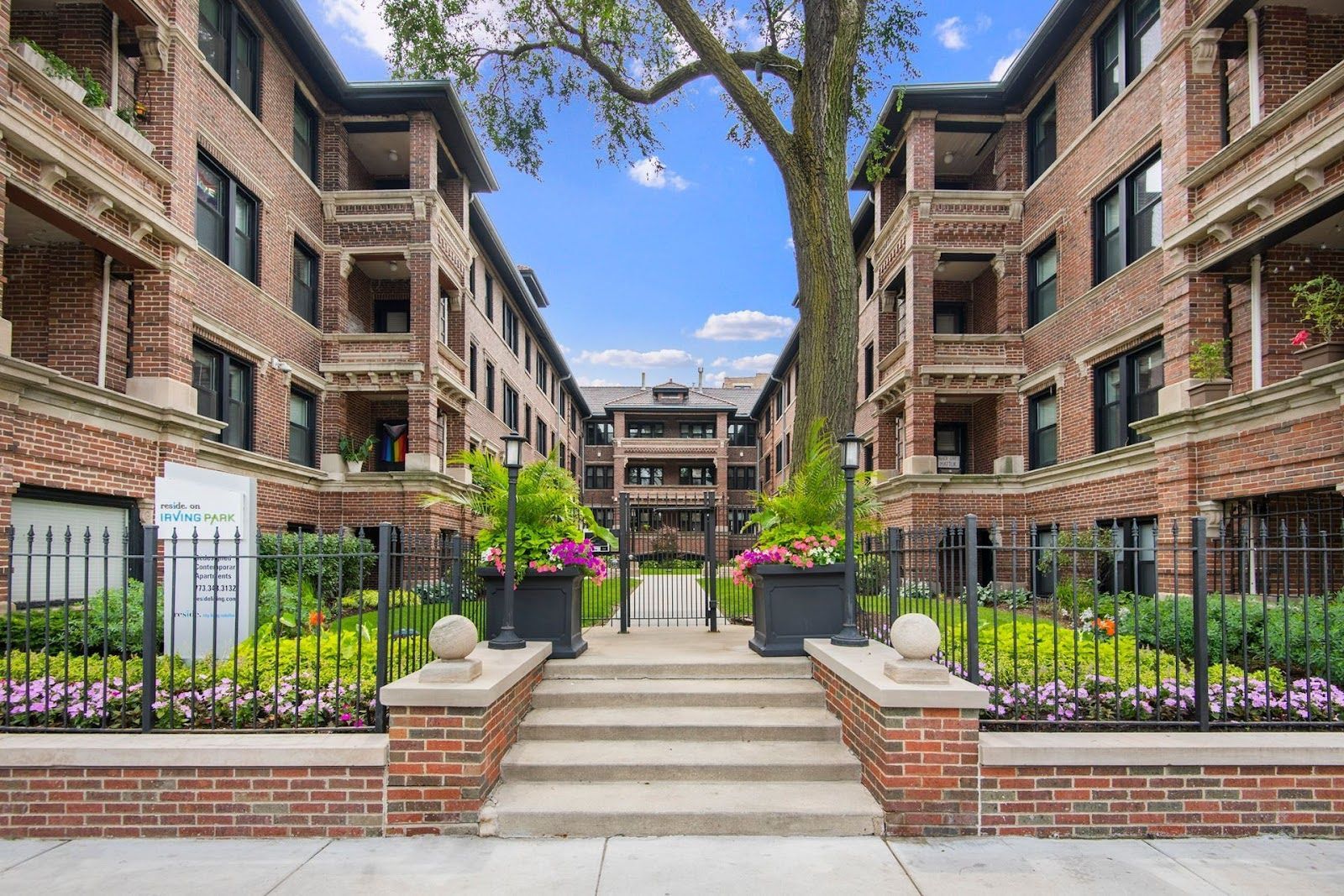 A brick apartment complex with a central courtyard, stone steps, metal fencing, and decorative planters.