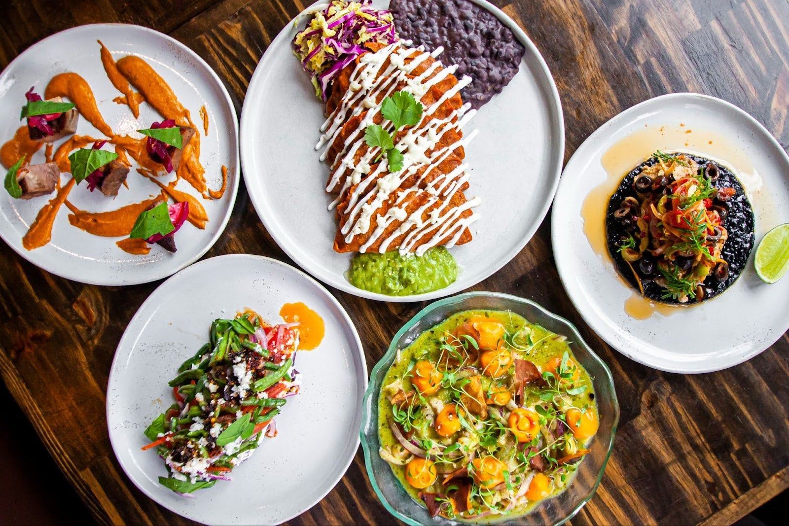 Five plates of colorful, artistically arranged gourmet Mexican-inspired dishes spread across a wooden table.