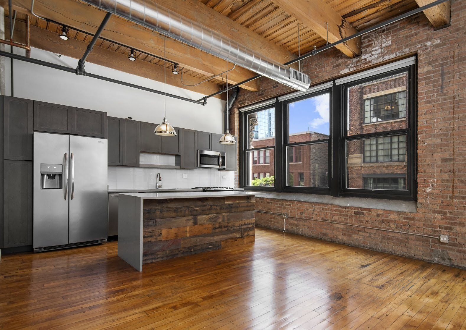 A modern kitchen in an industrial-style loft featuring exposed brick walls, wooden ceiling beams, and a wood-paneled island.