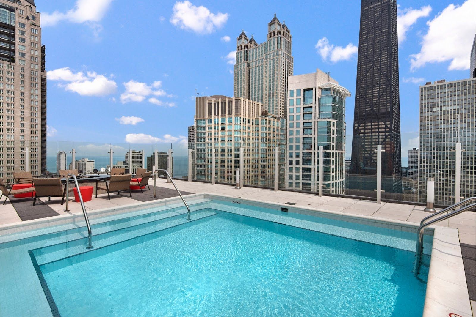 A rooftop swimming pool overlooking a city skyline with tall buildings under a blue, partly cloudy sky.