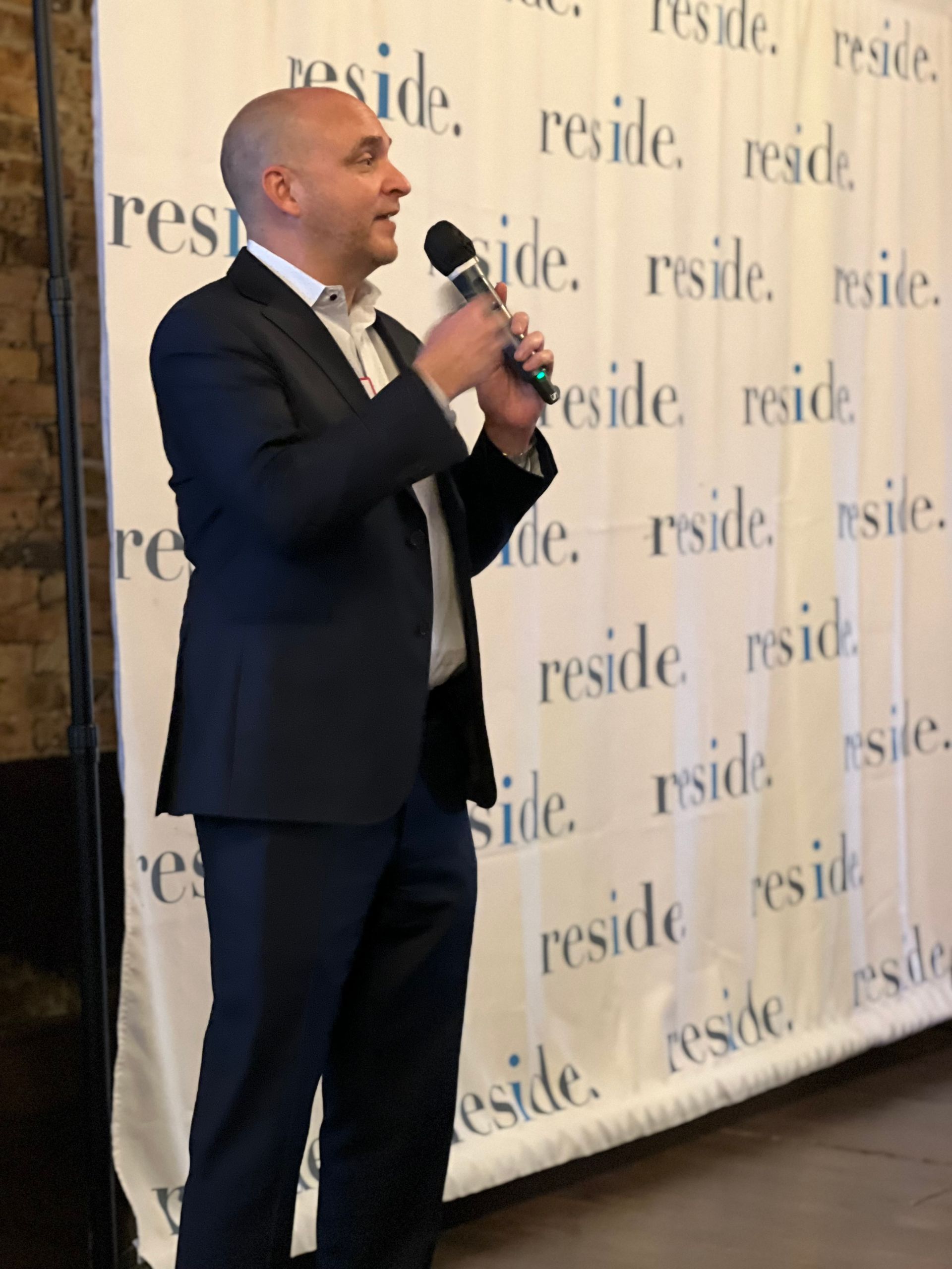 A man in a suit is holding a microphone in front of a wall that says reside