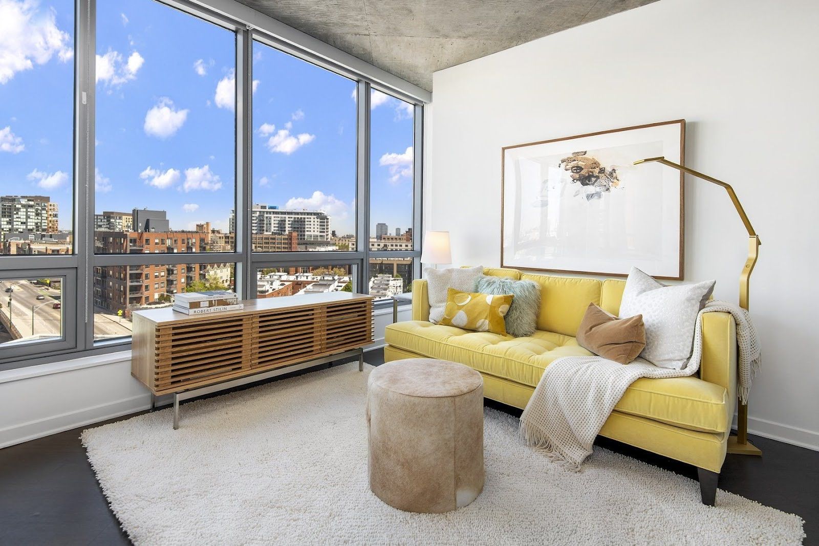 A modern living room with a yellow sofa, a textured wooden console, a circular ottoman, and a tall gold lamp by a window.