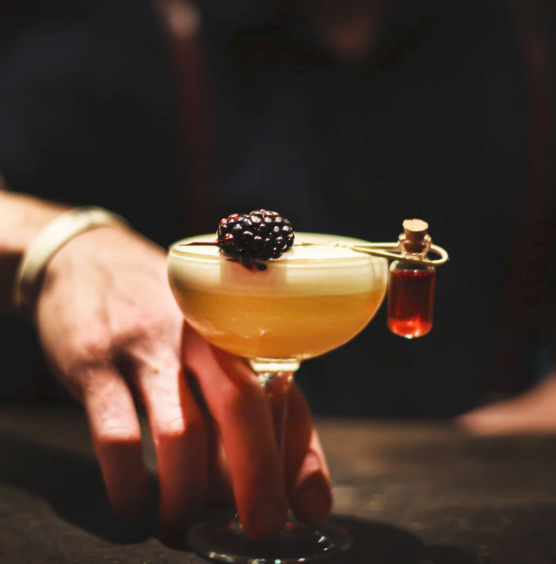 A person holds a coupe glass with a cocktail garnished with a blackberry and a miniature vial of liquid hanging from the rim.