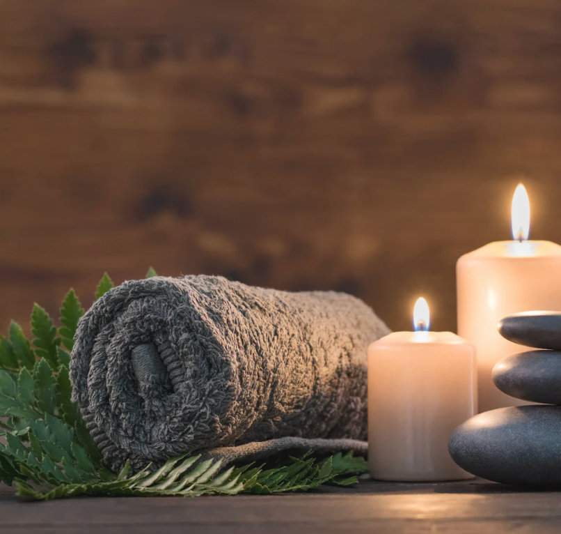Rolled gray towel, lit candles, and smooth stones on wood surface; spa setting.