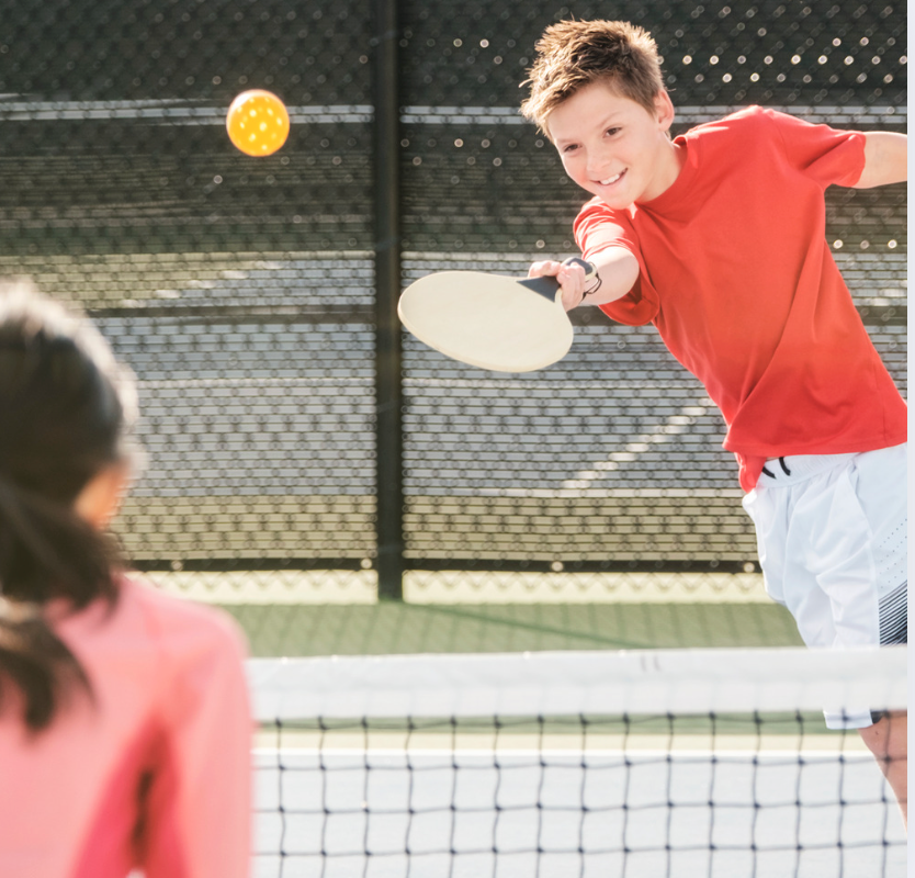 Boy in red shirt hitting a pickleball with a paddle, playing against a girl.