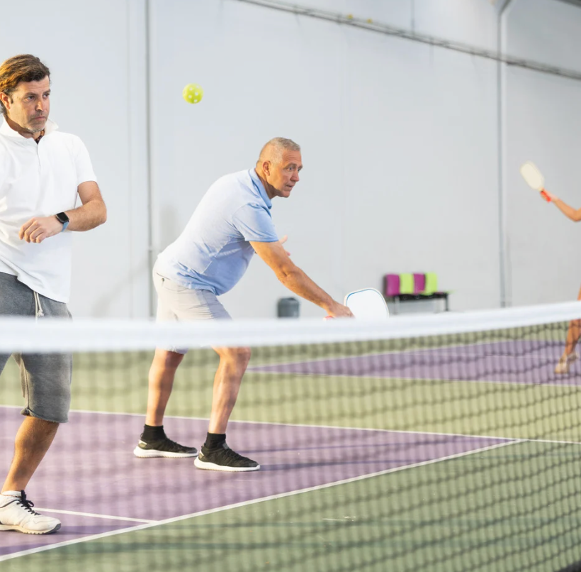 Two men playing pickleball indoors; one ready to hit ball, other watches. Net in foreground.