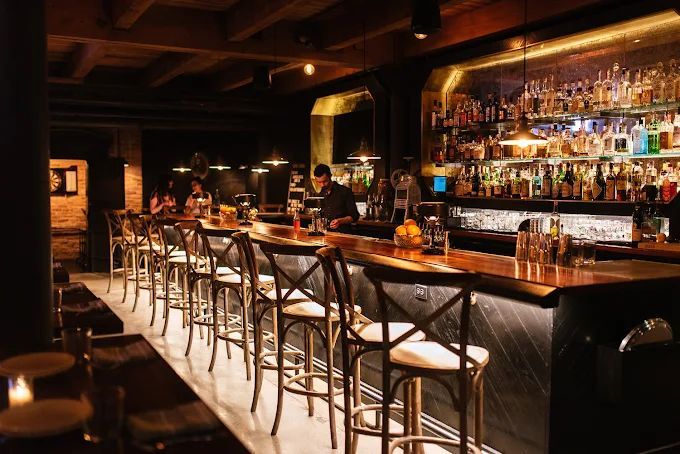 Dimly lit bar with a long wooden counter, rows of stools, and shelves filled with illuminated bottles behind the counter.