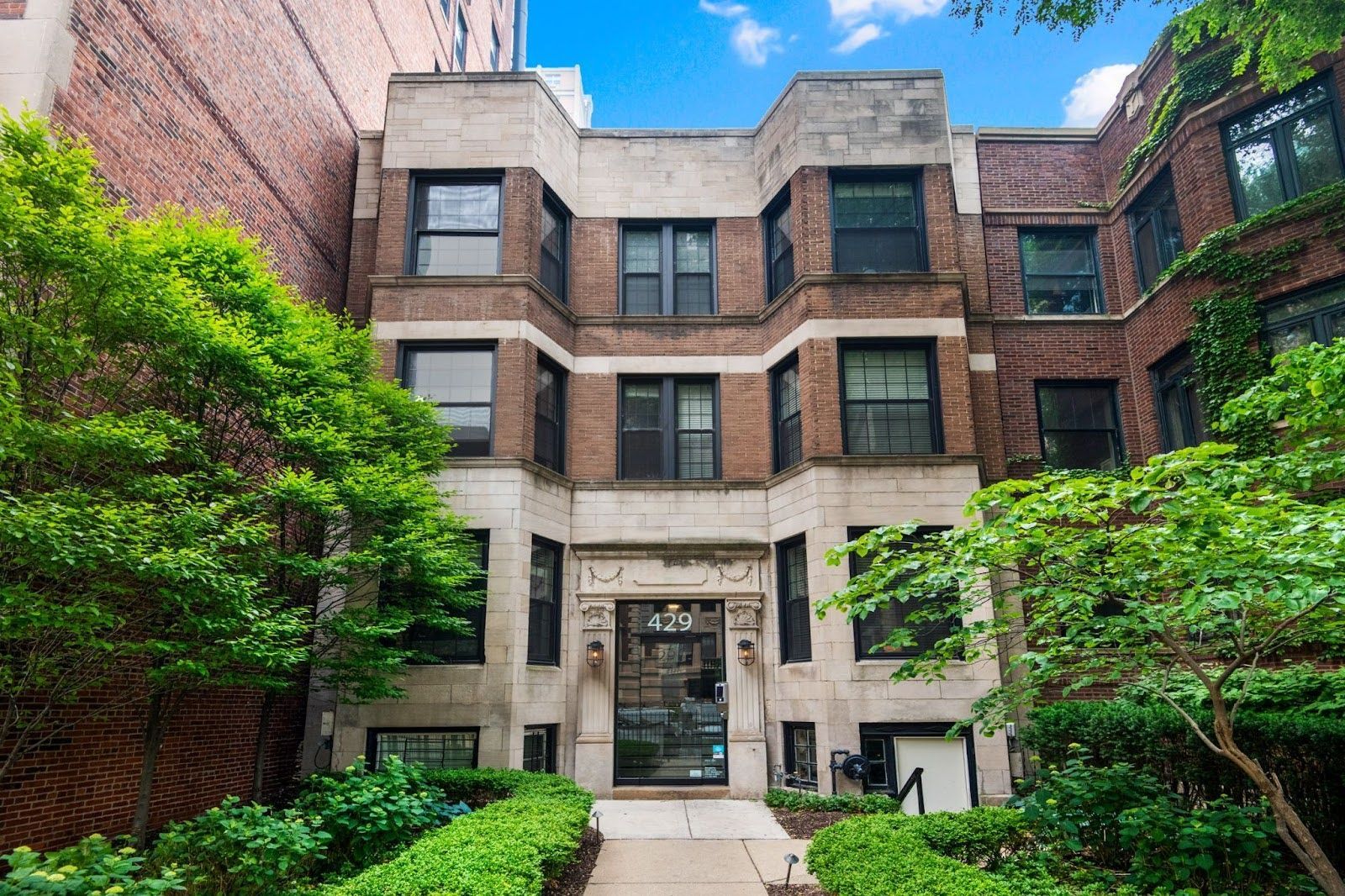 A brick apartment building with limestone accents, surrounded by greenery and a front walkway leading to the entrance.