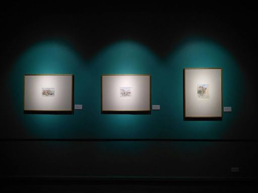 Three paintings are hanging on a green wall in a dark room.