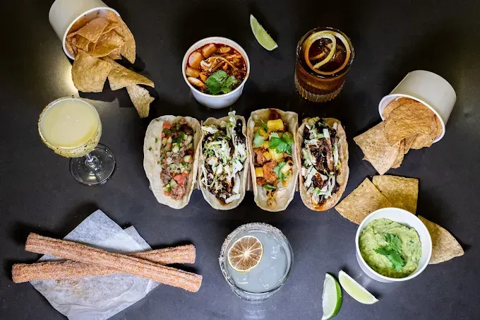 A flat-lay view of four tacos, guacamole, salsa, tortilla chips, churros, and three cocktails on a dark surface.