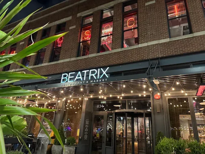 The storefront of Beatrix restaurant at night, featuring brick architecture, neon lights, and string lighting.