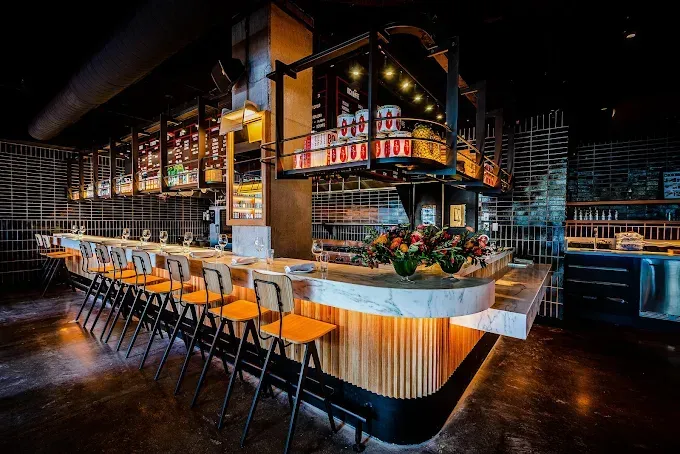 Modern, curved bar with wooden accents and light-colored countertops, featuring a row of chairs and illuminated shelving.