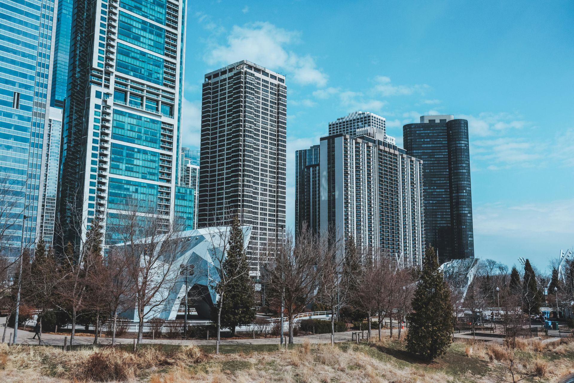 City skyline with tall buildings against a blue sky, trees in the foreground, and a modern art structure on the left.