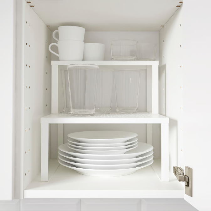 A white kitchen cabinet with two white shelf inserts, organizing stacked plates, clear glassware, and white mugs.