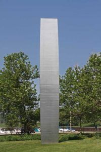 A tall stainless steel sculpture in a park with trees in the background.